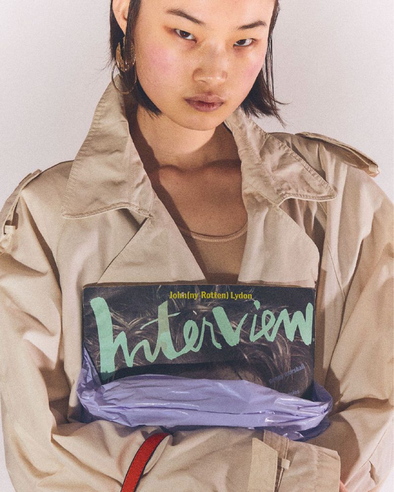 Asian woman in a trench coat holds Interview magazine with Johnny Rotten. Close-up studio portrait against a neutral backdrop.