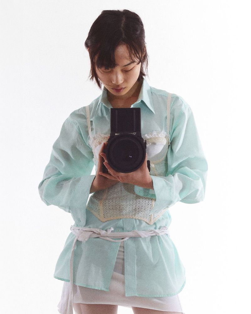 Young Asian woman with dark hair stands indoors holding a large-lens digital camera in front of a white background in eye-level shot.