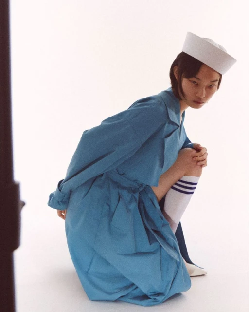 Young adult woman in a sailor hat and blue dress kneels in a photo studio wearing high socks with stripes, against a white backdrop.