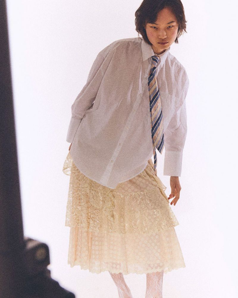 Adult woman in an oversized white dress shirt with a blue striped tie and lace skirt, posing in front of a bright white backdrop.