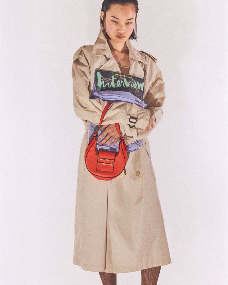 Asian woman in a trench coat holds a red purse against a white background in a fashion studio setting for a modern, stylish editorial.