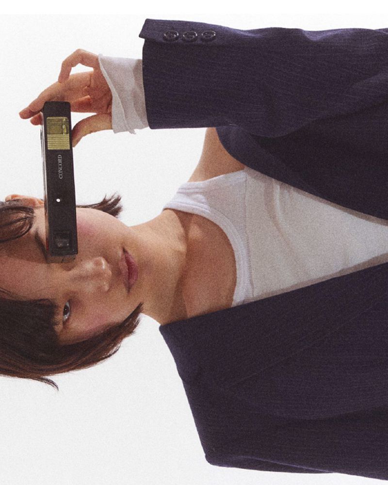 Portrait of a young person wearing a white top and a navy blazer, holding a black VHS tape to their face in a studio setting.