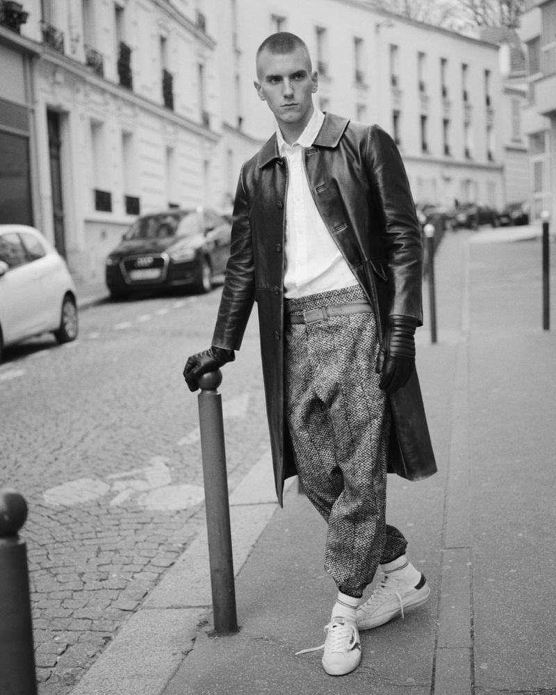 Monochrome shot of a man in a leather coat posing on a cobblestone street next to a bollard, with parked cars and buildings in the background.