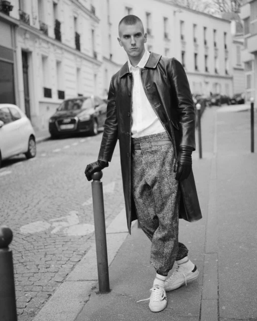 Monochrome shot of a man in a leather coat posing on a cobblestone street next to a bollard, with parked cars and buildings in the background.