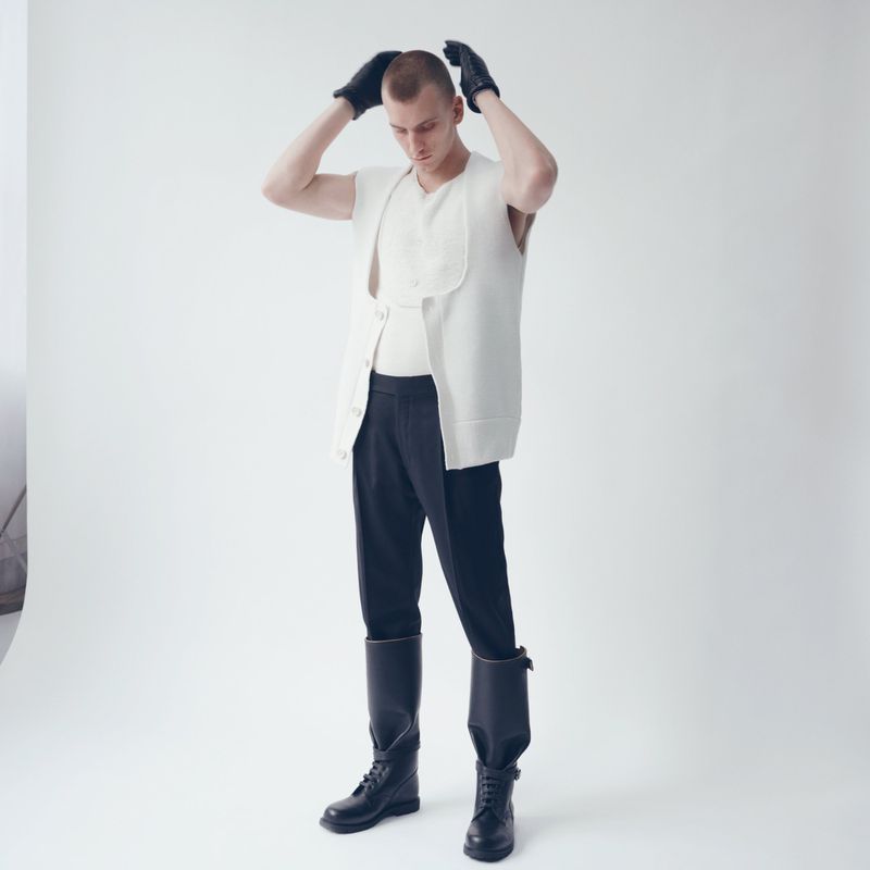 Bald man in cream vest and black gloves poses in a photo studio with a white background. He wears black pants and black boots.