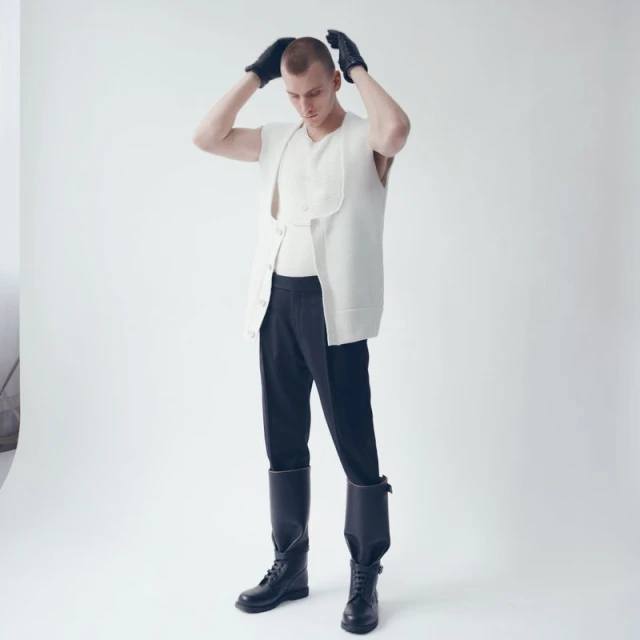 Bald man in cream vest and black gloves poses in a photo studio with a white background. He wears black pants and black boots.