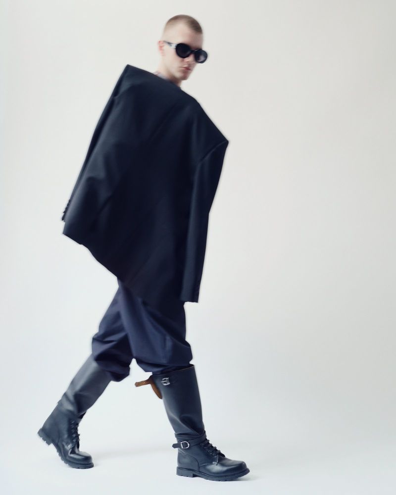 Man in a dark, oversized jacket, blue pants, and black boots walks against a white backdrop, wearing dark sunglasses, fashion shoot.