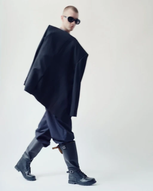 Man in a dark, oversized jacket, blue pants, and black boots walks against a white backdrop, wearing dark sunglasses, fashion shoot.