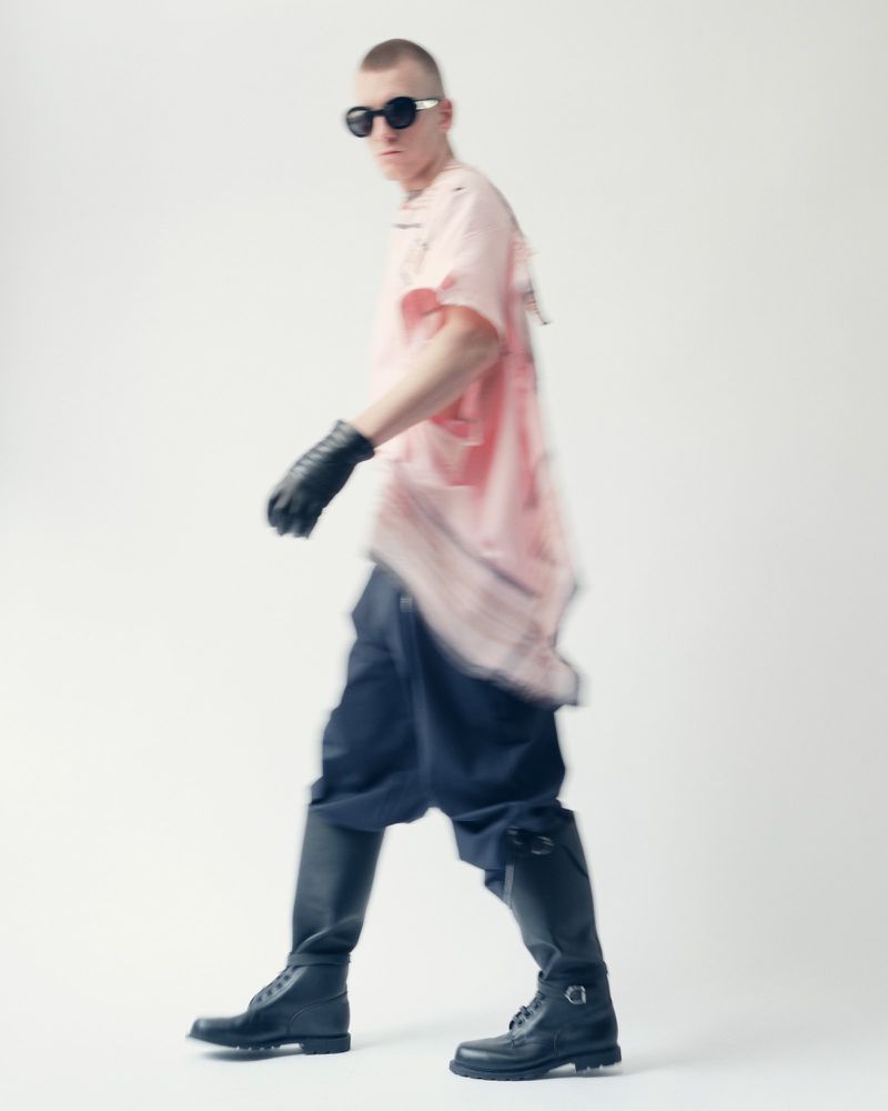 A person in black boots and sunglasses walks against a white backdrop, wearing a pink tee shirt and black gloves for a fashion shoot.