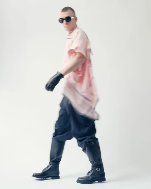 A person in black boots and sunglasses walks against a white backdrop, wearing a pink tee shirt and black gloves for a fashion shoot.