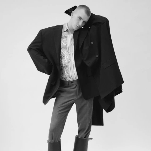 Monochrome studio shot of a fair-skinned adult man in oversized black blazer, patterned shirt, grey pants, and black boots against white.