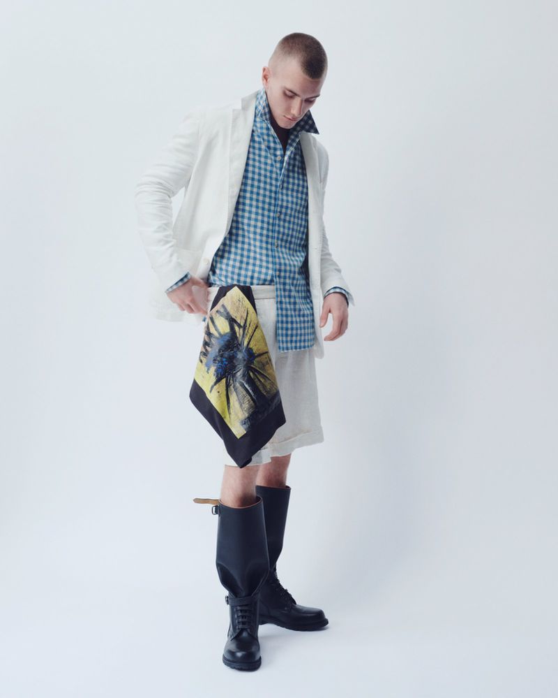 Male model in a white blazer and shorts, paired with a blue checkered shirt and black boots, stands in a bright, neutral photo studio.