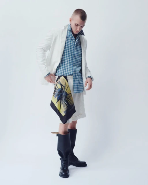 Male model in a white blazer and shorts, paired with a blue checkered shirt and black boots, stands in a bright, neutral photo studio.