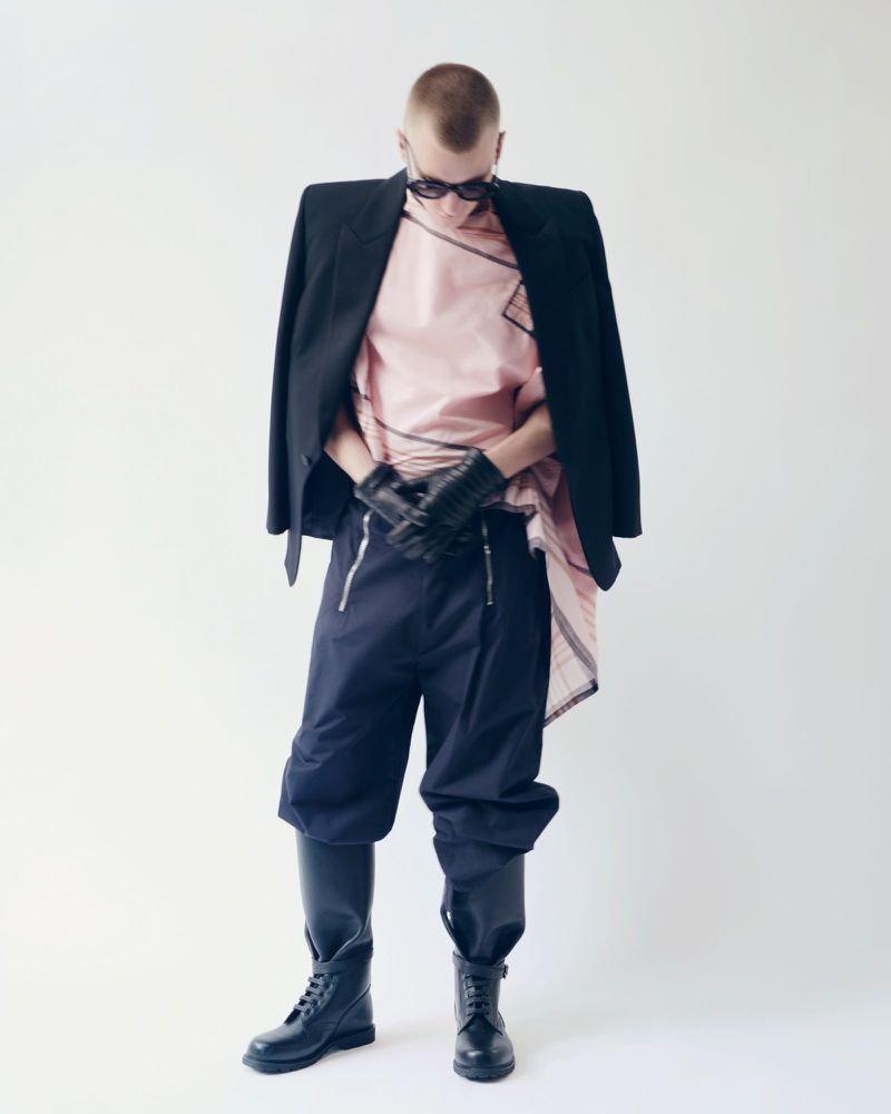 Adult male with shaved head poses in a pink top, black jacket, navy culottes, and black boots, standing against a plain white backdrop.