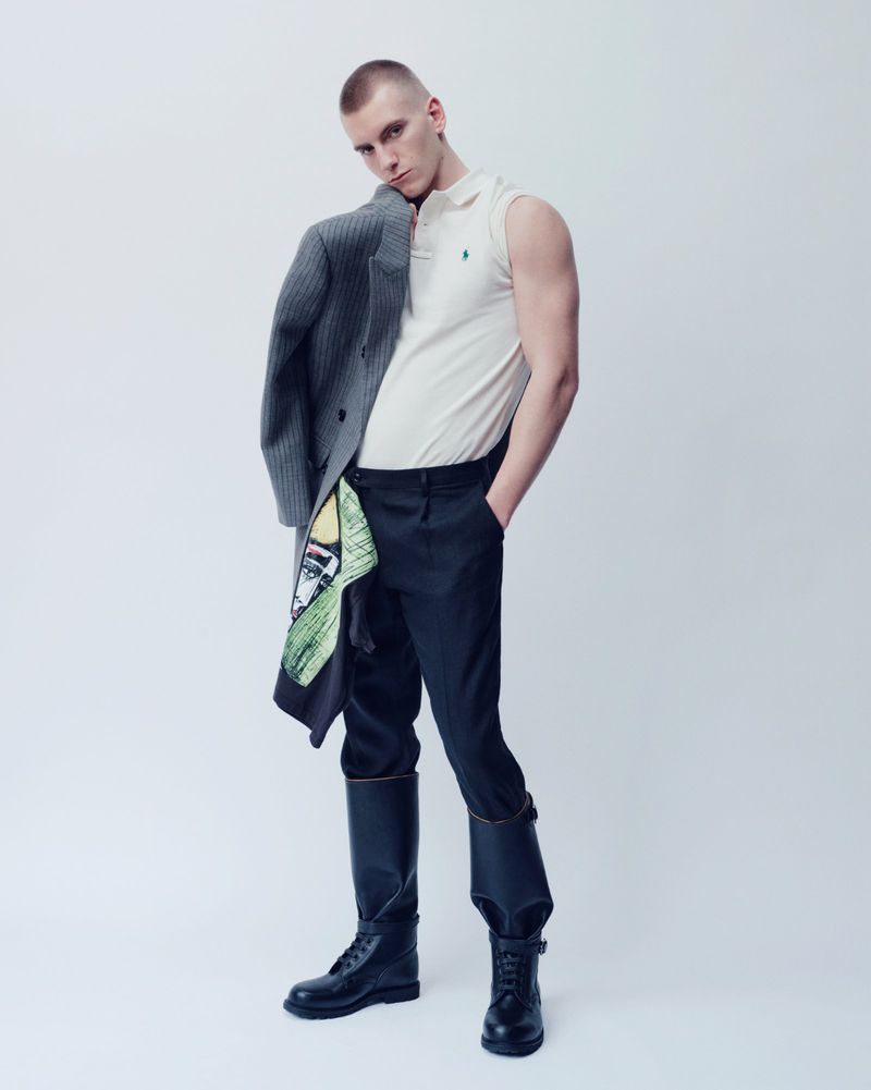 Man wearing a sleeveless white polo shirt and dark boots holding a grey coat over his shoulder
