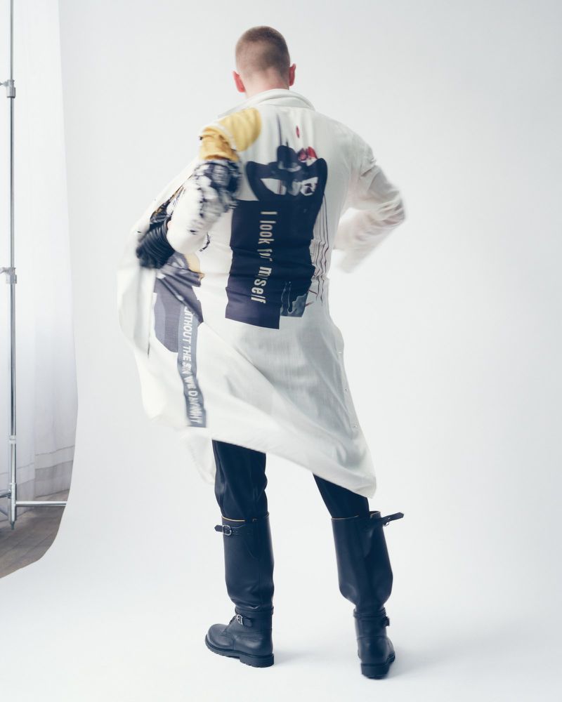 A slender adult male is shown from the rear, wearing a graphic-printed white jacket, dark pants, and black boots, in a photo studio setting.