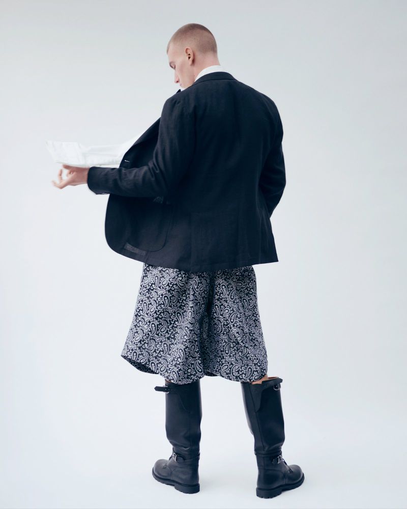 Back view of a fair-skinned man in a dark blazer, patterned shorts, and knee-high boots, holding a document in a bright photo studio.