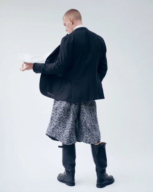 Back view of a fair-skinned man in a dark blazer, patterned shorts, and knee-high boots, holding a document in a bright photo studio.