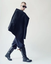 Model wearing dark sunglasses, oversized black blazer draped over shoulders, dark trousers, and black tall boots against a plain white background