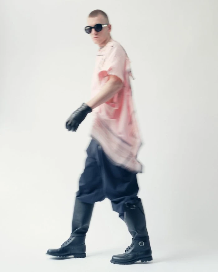 Model in dark sunglasses, pink shirt, black trousers, and tall boots walking against a plain white studio background.