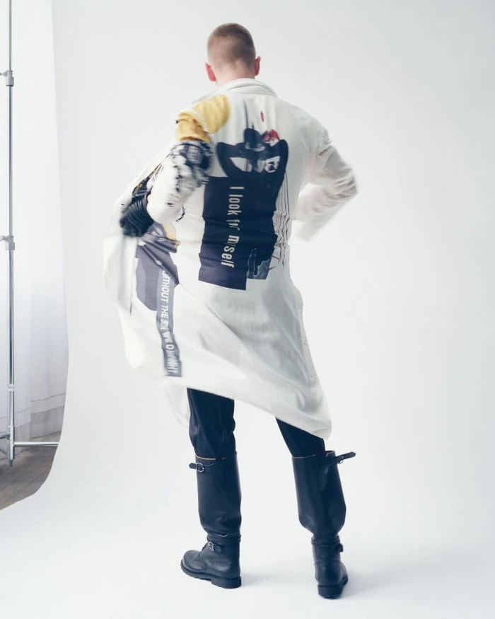 Model standing with back turned wearing a white printed coat and tall black boots against a white studio background