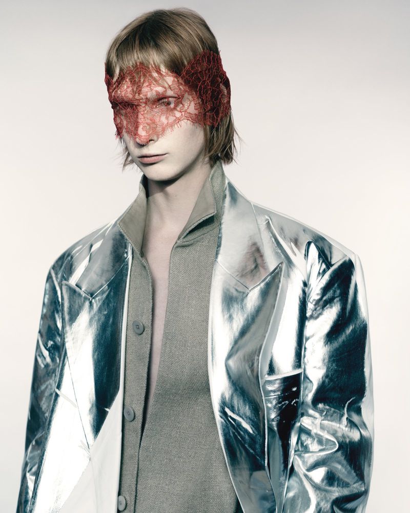 Close-up of a person with a red lace mask, wearing a grey top and transparent silver jacket against a neutral backdrop in a studio.