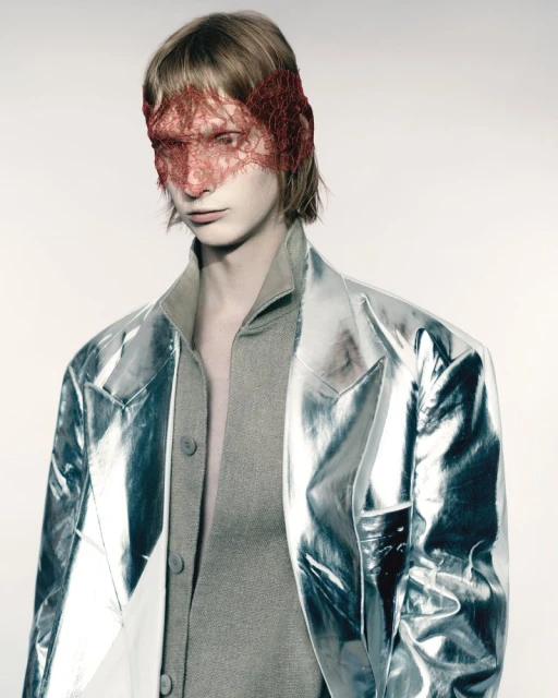 Close-up of a person with a red lace mask, wearing a grey top and transparent silver jacket against a neutral backdrop in a studio.