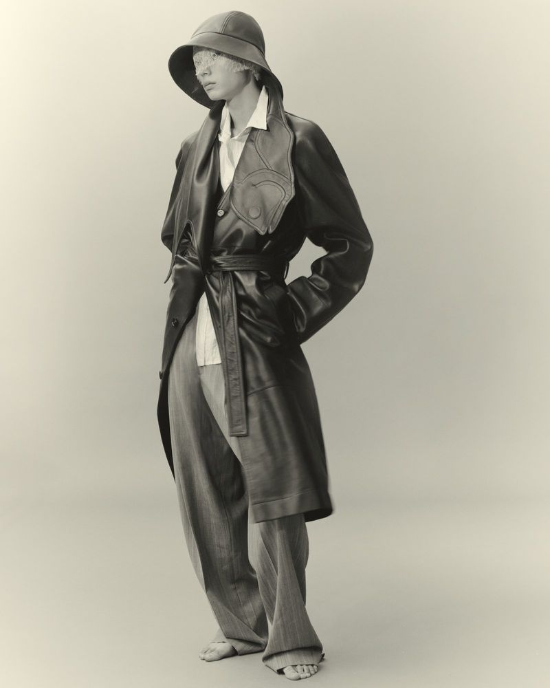 Full shot of person in oversized grey pants, button-down shirt, leather trench coat with tie, and brimmed hat against neutral background.