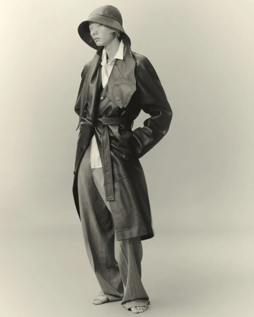 Full shot of person in oversized grey pants, button-down shirt, leather trench coat with tie, and brimmed hat against neutral background.