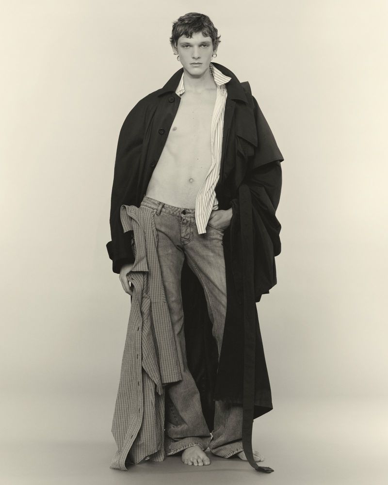 Model in dark coat and open striped shirt standing against plain background for U Repubblica project Ensemble