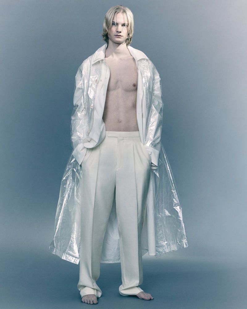Full-length studio shot of a bare-chested adult male model with long blonde hair, wearing wide-leg pants and a transparent shirt.