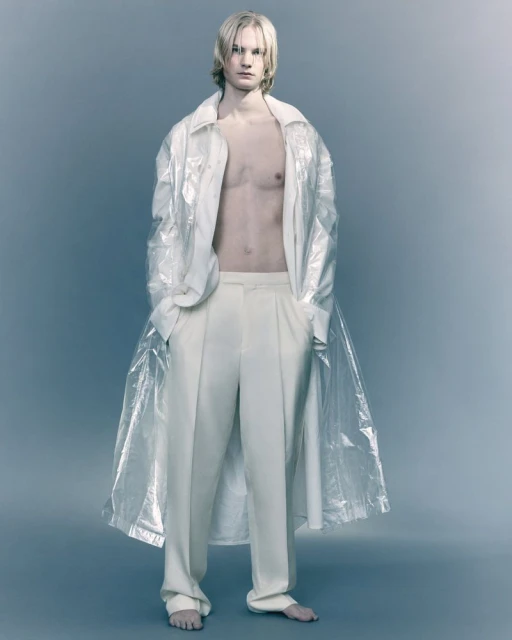 Full-length studio shot of a bare-chested adult male model with long blonde hair, wearing wide-leg pants and a transparent shirt.