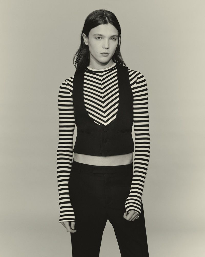 Studio shot of androgynous fashion model in black vest and striped top against neutral backdrop in a medium-shot portrait.