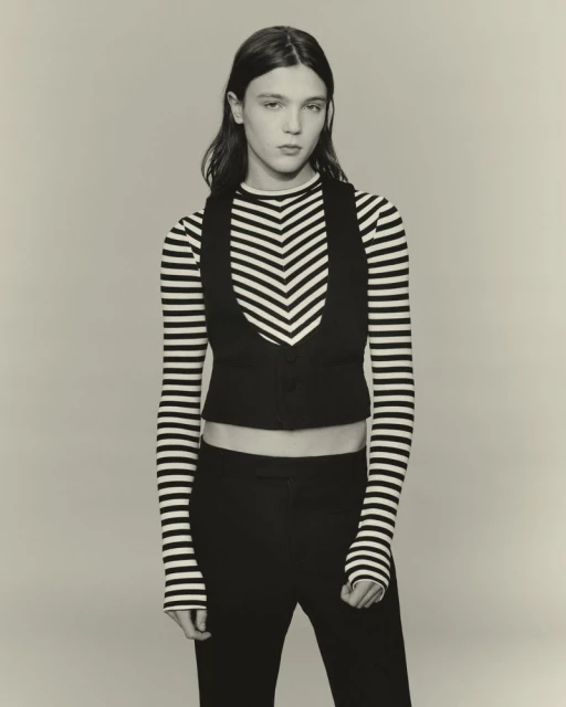 Studio shot of androgynous fashion model in black vest and striped top against neutral backdrop in a medium-shot portrait.
