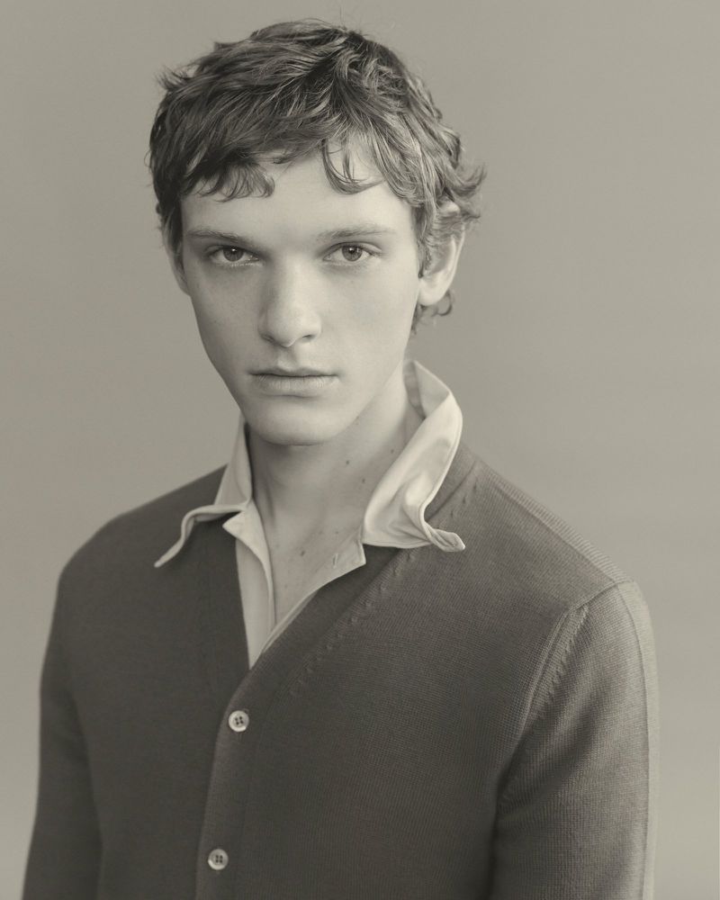 Portrait of a male model wearing a dark buttoned cardigan over a light collared shirt against a neutral grey studio background.