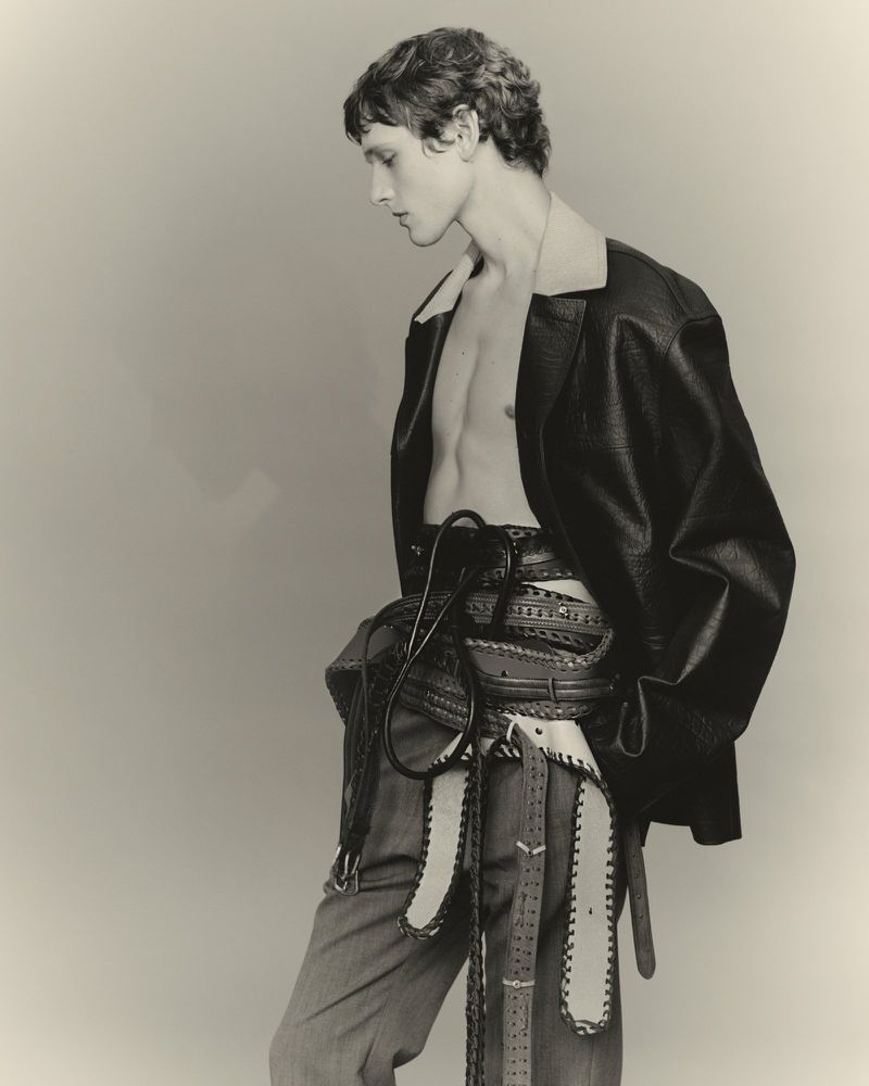 Male model in a black jacket and gray pants with decorative belts, striking a pose against a plain background in a fashion editorial photo.