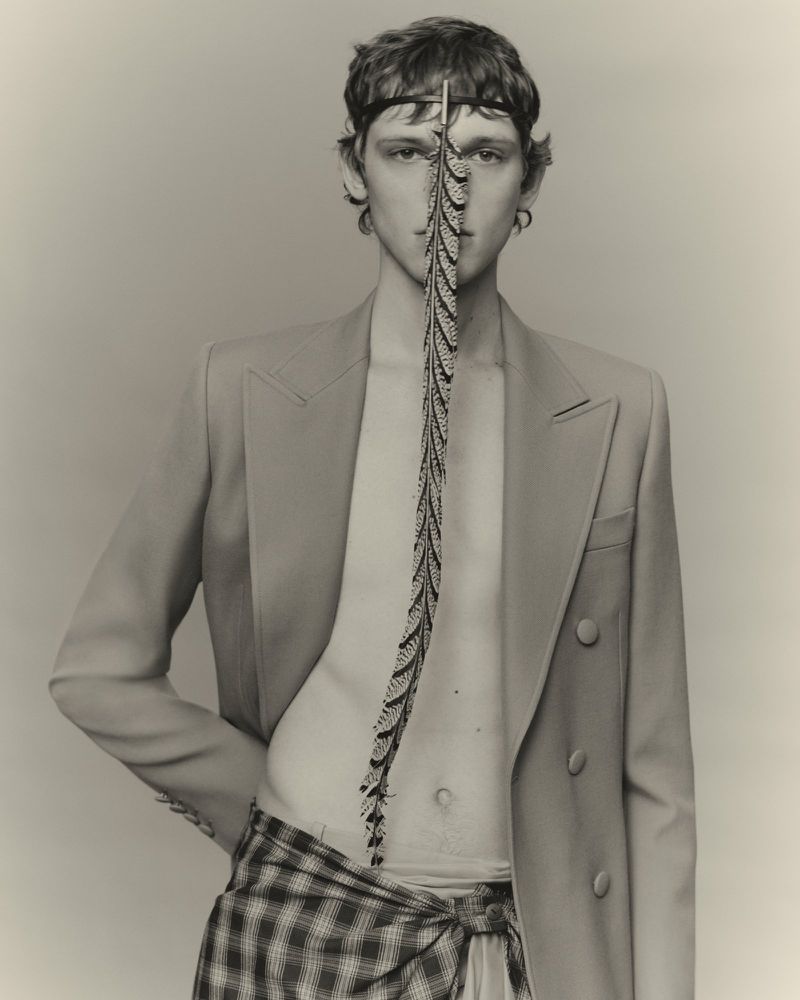 Portrait of a fair-skinned young male fashion model wearing a beige blazer and plaid skirt-wrap, styled with a feathered accessory.