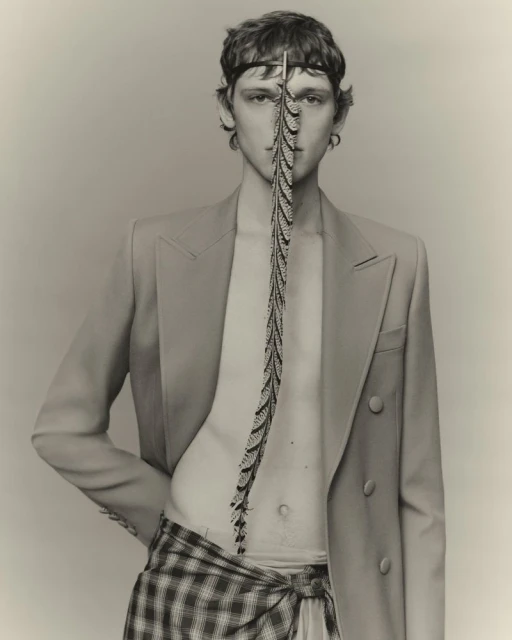 Portrait of a fair-skinned young male fashion model wearing a beige blazer and plaid skirt-wrap, styled with a feathered accessory.