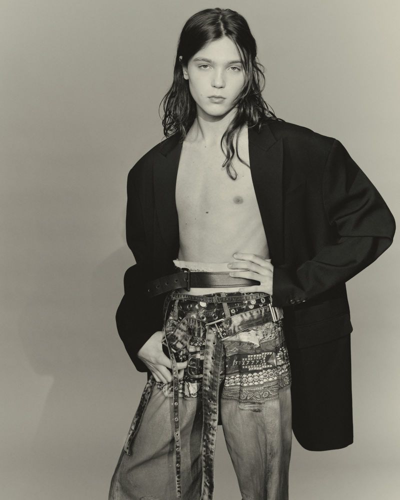 Adult male with long hair poses in a studio wearing an oversized black blazer and patterned denim pants with a belt. Fashion.