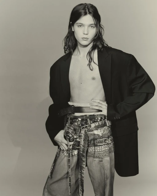 Adult male with long hair poses in a studio wearing an oversized black blazer and patterned denim pants with a belt. Fashion.