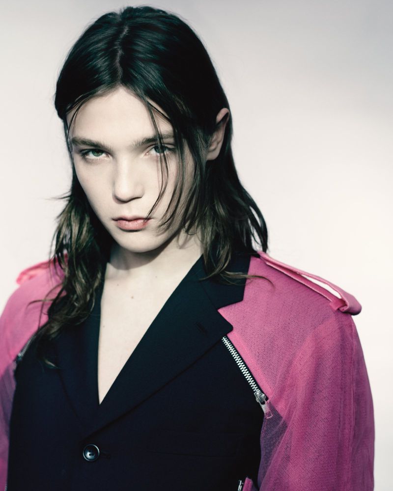 Portrait of a young adult with long, dark hair wearing a pink and black jacket against a pale background, captured in a studio setting.