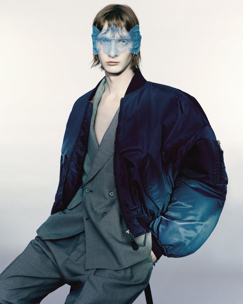 Portrait of a slender adult male wearing a blue mask and a gray suit with a blue bomber jacket, posing indoors against a white background.