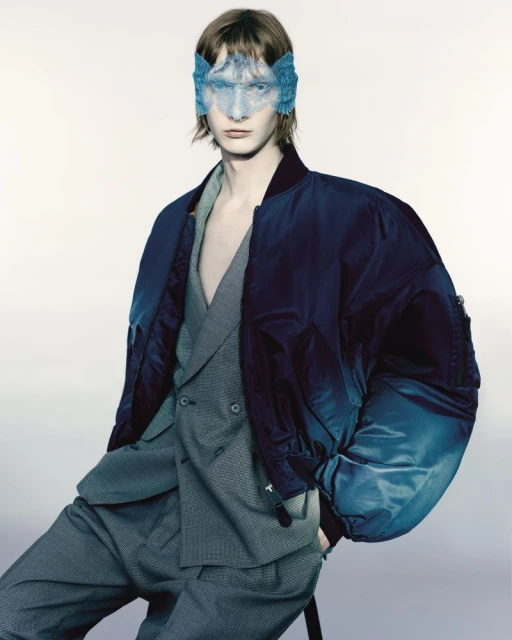 Portrait of a slender adult male wearing a blue mask and a gray suit with a blue bomber jacket, posing indoors against a white background.