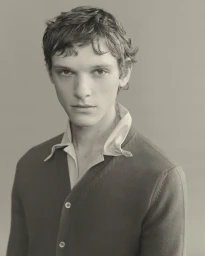 Portrait of a male model wearing a dark buttoned cardigan over a light collared shirt against a neutral grey studio background.