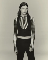 Model wearing a black vest over a striped long-sleeve top against a neutral backdrop in an editorial portrait