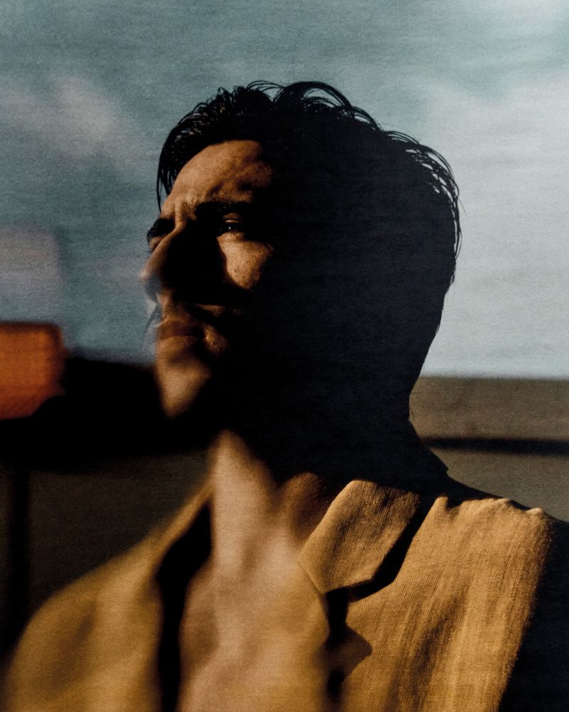 Headshot of a man in a jacket looking to the left, with dark hair, angular jawline, and sky and shadows in the background.
