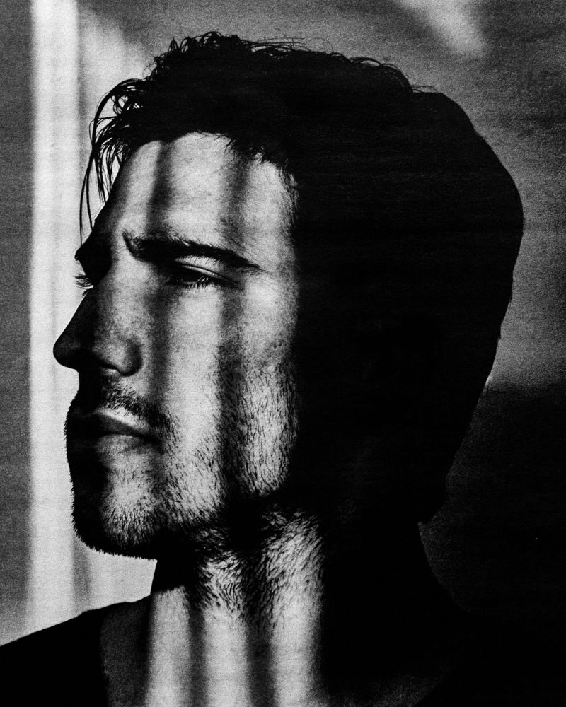 Black and white portrait of an adult male with strong shadows creating stripes across the face, wearing a dark top.