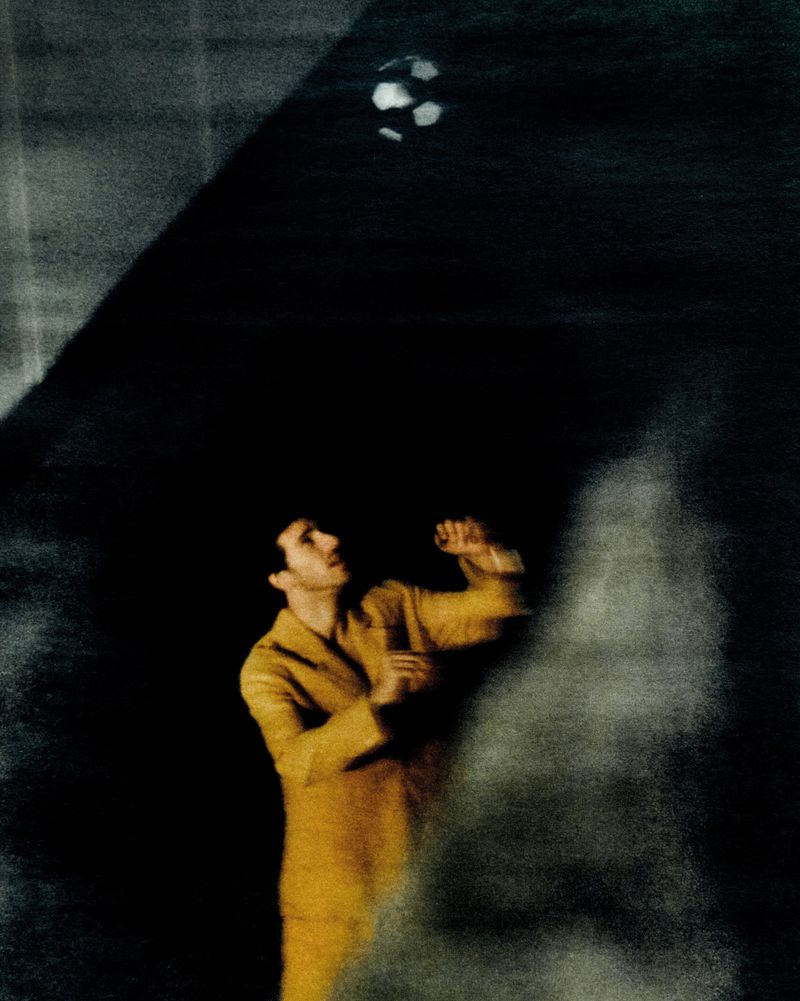 Man in mustard jacket reaches for soccer ball in dark, shadowed outdoor space with blurred, dreamlike quality and limited color palette.