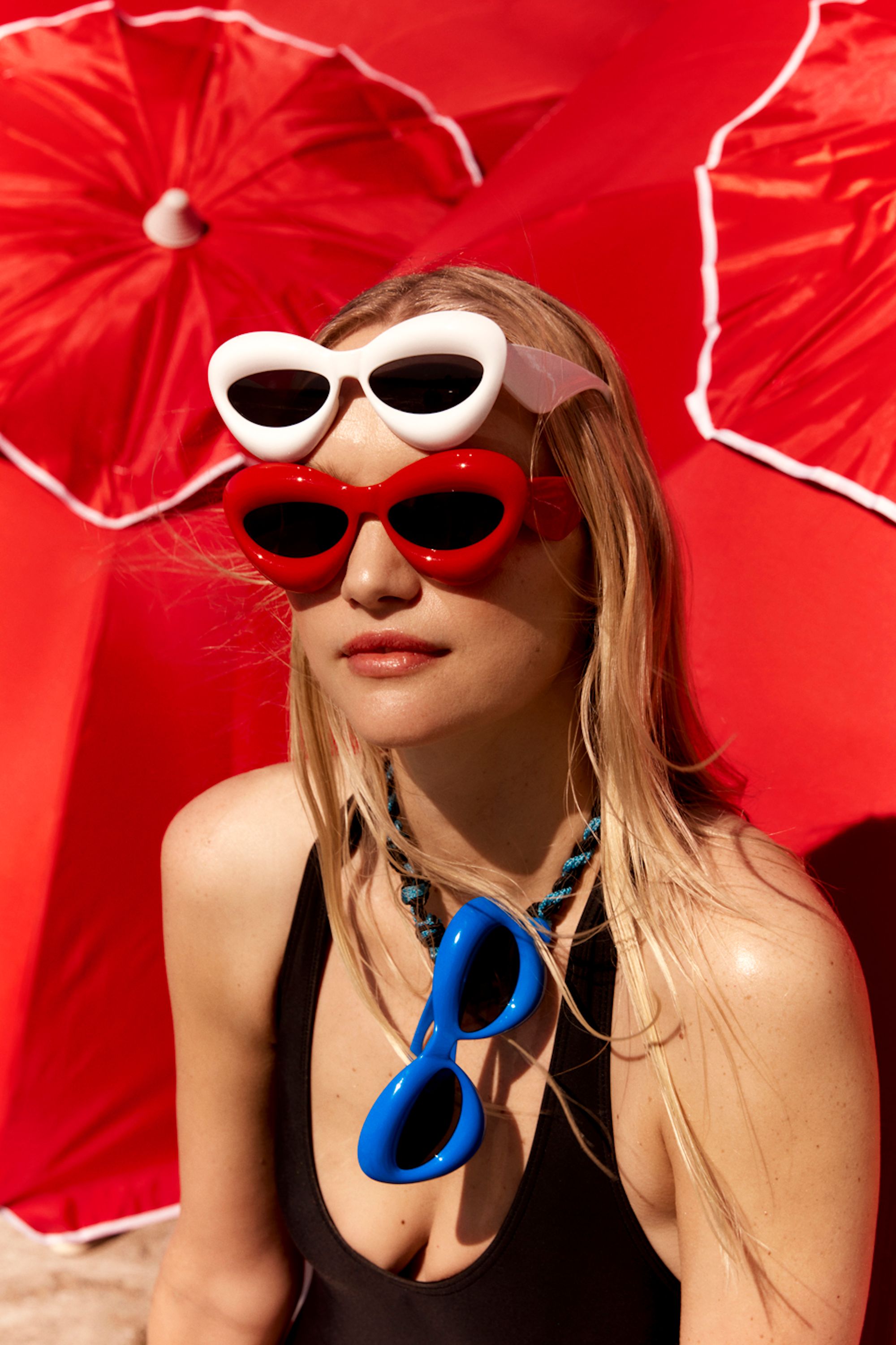 Woman wears red and white sunglasses with blue glasses necklace against red umbrella backdrop in eye-level medium shot.