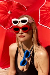 Close-up portrait of a blonde woman wearing red sunglasses, holding white sunglasses on her forehead with red umbrellas behind.